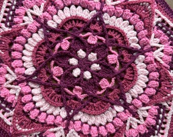 May include: A handmade, crocheted blanket in a floral design. The blanket features a starburst pattern with shades of pink, white, and deep purple. The crochet work is detailed, with raised textures and intricate stitch patterns. The blanket is a square shape.