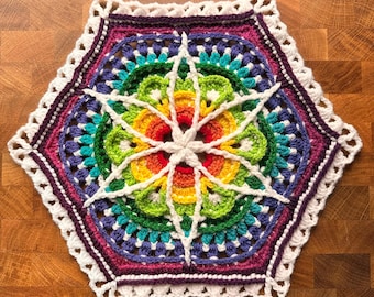 May include: A colorful, handmade crochet doily with a hexagonal shape. The design features concentric circles of vibrant colors, including red, orange, yellow, green, blue, and purple. A white starburst pattern is at the center, and the edges are finished with a scalloped border.