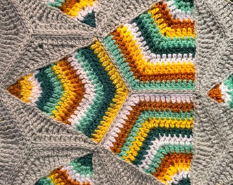 May include: A crochet blanket featuring a geometric pattern of repeating star-shaped motifs. Each motif is composed of concentric layers in shades of gray, white, yellow, dark green, light green, and brown, joined by solid gray yarn.