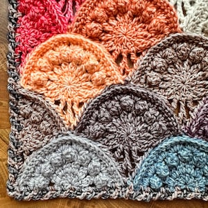 May include: A close-up of a crocheted blanket with a variety of colors, including pink, orange, brown, gray, and blue. The blanket is made up of a series of circular motifs that are joined together.