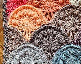 May include: A close-up of a crocheted blanket with a variety of colors, including pink, orange, brown, gray, and blue. The blanket is made up of a series of circular motifs that are joined together.