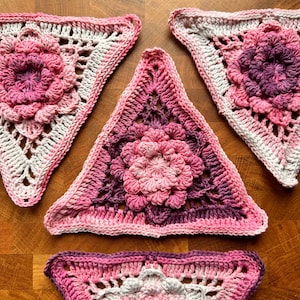 Puede incluir: Conjunto de piezas de ganchillo hechas a mano en tonos rosas y morados. Las piezas son triangulares y presentan un diseño floral central. El trabajo de ganchillo es detallado, con patrones calados y bordes texturizados. Se exhiben sobre una superficie de madera.