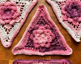 May include: A collection of handmade crochet pieces in shades of pink and purple. The pieces are triangular and feature a central floral design. The crochet work is detailed, with openwork patterns and textured edges. The items are displayed on a wooden surface.