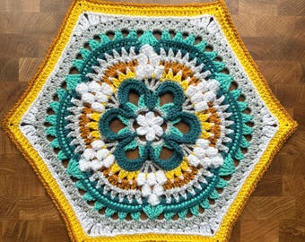 May include: A handmade, colorful crochet doily in a hexagonal shape. The design features a central flower motif in white and teal, surrounded by concentric rings of teal, yellow, gray, and brown. The outer edge is bordered with yellow and brown.