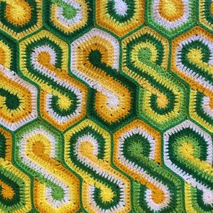 Op de afbeelding: Een gehaakt deken met een geometrisch patroon in tinten groen, geel en wit. Het deken is gemaakt van zacht, getextureerd garen en heeft een honingraatontwerp.
