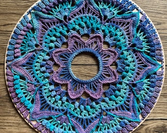 May include: A crocheted mandala wall hanging with a circular design in shades of purple, blue, and green. The mandala is made with a variety of stitches and textures, creating a visually interesting and intricate pattern.