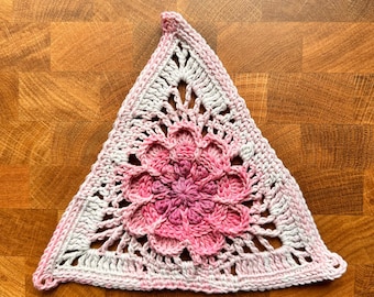 May include: A handmade, triangular crochet piece featuring a central floral design. The piece is primarily white with a pink and white gradient flower in the center. The edges are finished with a pink border, creating a decorative and textured item.