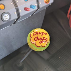 Könnte beinhalten: Ein individueller Schaltknauf mit dem Chupa Chups-Logo. Der Knopf ist gelb mit einem grünen Rand und dem Chupa Chups-Text in Rot. Der silberne Schalthebel ist vor dem Fahrzeuginnenraum zu sehen.