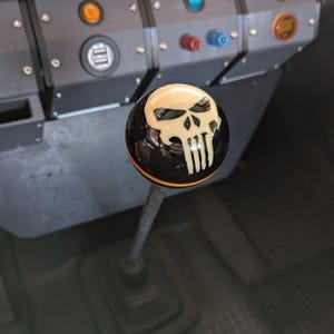 May include: A black and cream skull shift knob with a chrome shift lever. The skull design is a stylized, white graphic on a glossy black sphere. The knob is set against a dark interior.