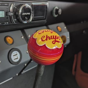 Könnte beinhalten: Ein individueller Schaltknauf mit einem Chupa Chups-Logo. Der Knauf ist ein rotes und gelbes Lollipop-Design mit orangefarbenen und rosa Streifen. Der Schalthebelknauf ist im Innenraum eines Fahrzeugs eingebaut.