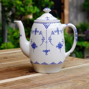 Pode incluir: Cafeteira de cerâmica branca com tampa, adornada com padrões florais e de videira azuis. A cafeteira tem um bico e uma alça curvos e está sobre uma mesa de madeira. O design inclui uma base e uma borda da tampa onduladas.