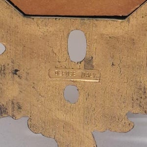 May include: Close-up of a gold-colored decorative metal piece with an ornate design. The piece has two oval-shaped holes and the text "DEPOSE ITALY" is visible.
