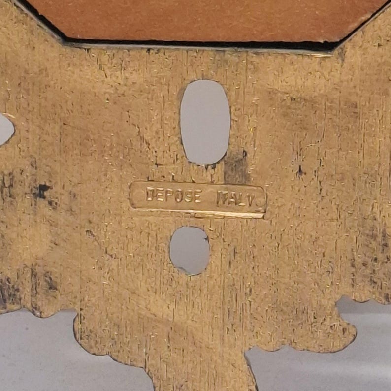 May include: Close-up of a gold-colored decorative metal piece with an ornate design. The piece has two oval-shaped holes and the text "DEPOSE ITALY" is visible.
