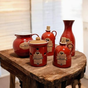 Op de afbeelding: Een verzameling rode keramische keukencontainers met kurken en etiketten. De set bevat een waterkan, een knoflookkelder, twee olieflessen en een hoge vaas. De containers staan op een rustieke houten tafel.