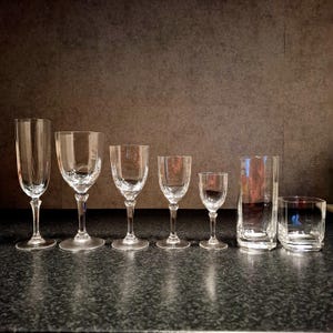 May include: A collection of seven clear glass drinkware items, including champagne flutes, wine glasses, and tumblers. The glasses are arranged in a row, showcasing various shapes and sizes. The glassware is displayed on a dark surface.
