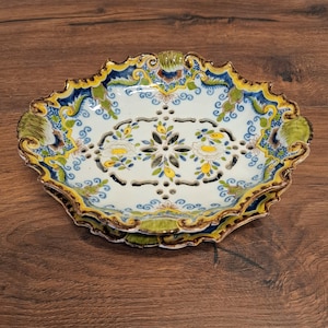 Op de afbeelding: Een ovale keramische schaal met een opengewerkt ontwerp. De schaal heeft een witte basis met bloemen en geometrische patronen in geel, blauw en groen. De geschulpte rand is afgewerkt met goud. Een decoratief stuk om te serveren of tentoon te stellen.