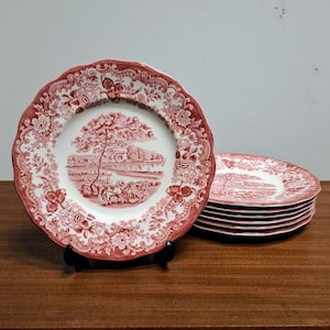 May include: A collection of antique ceramic plates featuring a red and white floral and landscape pattern. The plates have a scalloped edge and are stacked on a wooden surface. These plates are a classic style for serving food.