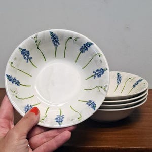 Op de afbeelding: Een stapel witte keramische kommen met een handgeschilderd patroon van blauwe druifhyacinten en groene stengels, gerangschikt in een cirkelvormig motief rond de binnenrand. De kommen hebben een gladde, glanzende afwerking en een ronde vorm.