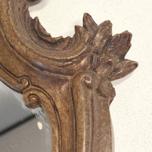 Puede incluir: Primer plano de un marco de espejo marr&oacute;n y ornamentado. El marco presenta tallas detalladas con un dise&ntilde;o floral en la parte superior y detalles en espiral. La superficie del espejo es visible dentro del marco, reflejando un fondo claro.