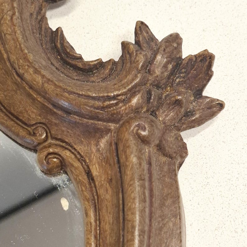 Puede incluir: Primer plano de un marco de espejo marr&oacute;n y ornamentado. El marco presenta tallas detalladas con un dise&ntilde;o floral en la parte superior y detalles en espiral. La superficie del espejo es visible dentro del marco, reflejando un fondo claro.