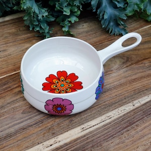 Op de afbeelding: Witte keramische kom met een handvat, versierd met een kleurrijk bloemenontwerp. De kom heeft een patroon van rode, oranje, gele, blauwe en roze bloemen.