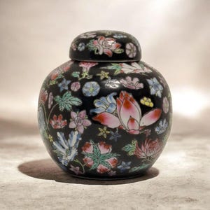 Pode incluir: Um jarro de cerâmica preto com tampa a condizer, adornado com um padrão floral vibrante em rosa, vermelho, azul e verde. O jarro tem uma forma arredondada e é provavelmente usado para fins decorativos.