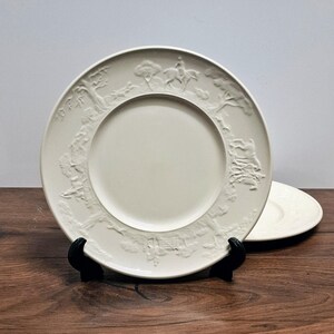 Op de afbeelding: Crème-witte keramische plaat met een reliëf ontwerp rond de rand. Het ontwerp toont een jachttafereel met bomen, een ruiter te paard en andere figuren. Een tweede plaat is zichtbaar op de achtergrond.