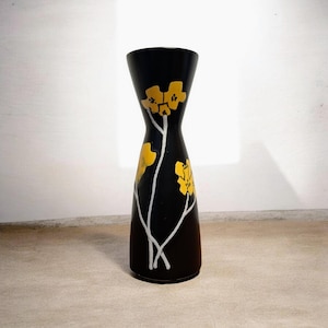 Könnte beinhalten: Eine schwarze Vase mit einer schlanken Sanduhrform. Die Vase hat gelbe Blumendesigns mit weißen Stielen. Die Vase ist etwa 25 cm hoch und eignet sich zum Präsentieren von Blumen oder als dekoratives Element.
