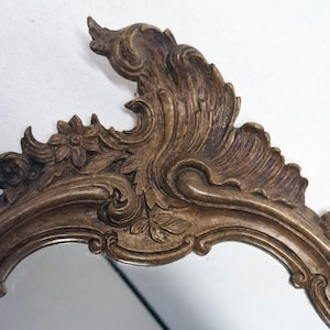 Puede incluir: Primer plano de un espejo decorativo con un marco ornamentado de estilo antiguo. El marco es de color bronce c&aacute;lido y presenta dise&ntilde;os detallados de flores y volutas. La forma del espejo es suave y redondeada, reflejando el entorno.