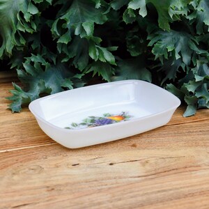 Peut inclure: Un plat rectangulaire blanc aux coins arrondis. Le plat présente un motif floral coloré avec des raisins violets et un fruit orange. Le fond comprend un feuillage vert, posé sur une surface en bois.