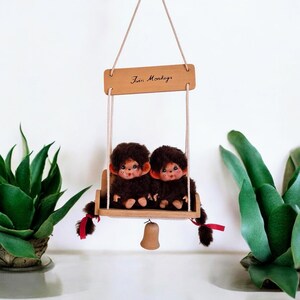 Può includere: Due scimmie di peluche marroni con fiocchi rossi sedute su un'altalena di legno. L'altalena è sospesa da una corda e ha un cartello di legno con la scritta "Twin Monkeys". Una piccola campana è appesa sotto.