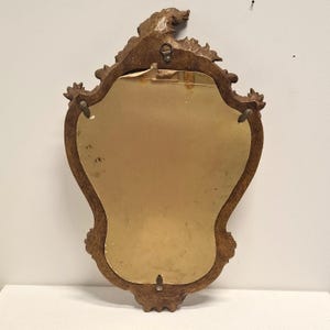 Puede incluir: Un espejo vintage con un marco de madera tallada marr&oacute;n. La parte posterior del espejo es visible, mostrando una superficie dorada y herrajes met&aacute;licos para colgar. El marco presenta detalles decorativos florales y de volutas.