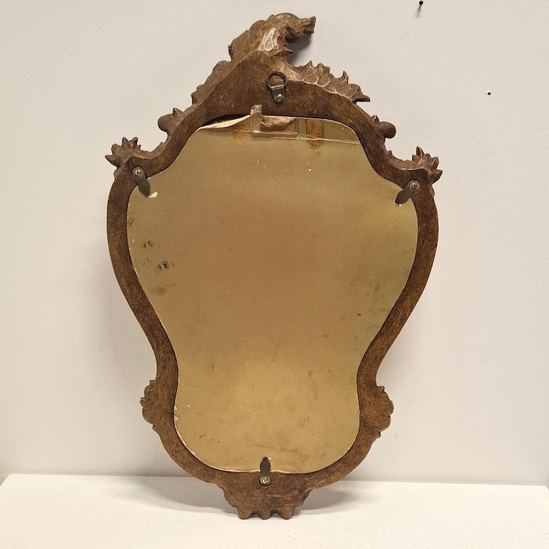 Puede incluir: Un espejo vintage con un marco de madera tallada marr&oacute;n. La parte posterior del espejo es visible, mostrando una superficie dorada y herrajes met&aacute;licos para colgar. El marco presenta detalles decorativos florales y de volutas.