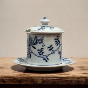 Op de afbeelding: Een witte en blauwe keramische suikerpot met deksel en een kleine lepel, rustend op een bijpassende schotel. De pot en schotel hebben een blauw bloemen- en wijnstokpatroon. De set wordt gepresenteerd op een houten oppervlak.