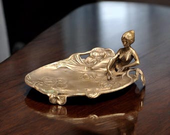 May include: A gold-toned Art Nouveau style dish with a reclining female figure. The dish has an oval shape with floral and leaf details. The figure is seated on the edge of the dish.