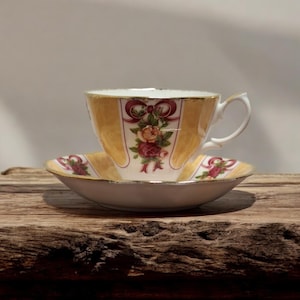 Op de afbeelding: Een vintage theekop en schotel set. De theekop en schotel zijn versierd met een geel en wit gestreept patroon, bordeauxrode strikken en een bloemenboeket. De set is gemaakt van porselein en heeft een gouden rand.