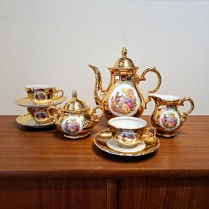 Puede incluir: Juego de té dorado vintage con tetera, azucarero, jarrita para la leche y dos tazas con platillos. Cada pieza presenta una escena pintada con figuras en un entorno de jardín. El juego se exhibe sobre una superficie de madera.