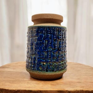 Pode incluir: Um vaso de cerâmica com exterior azul escuro texturizado e borda e base marrons. O vaso tem formato cilíndrico e assenta sobre uma superfície de madeira. O design apresenta um padrão repetitivo de pequenos quadrados em relevo.