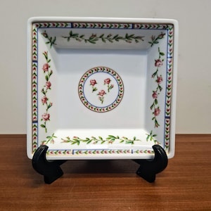 Op de afbeelding: Een vierkante witte porseleinen schaal met een centraal rond medaillon met drie roze rozenknoppen. De rand is versierd met een herhalend patroon van roze rozen, groene bladeren en blauwe lijnen. De schaal staat op een zwarte kunststof standaard.