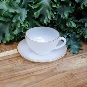 Könnte beinhalten: Eine weiße Teetasse und Untertasse mit goldener Verzierung. Tasse und Untertasse stehen auf einer Holzoberfläche.