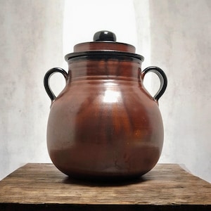 Könnte beinhalten: Ein brauner Keramiktopf mit Deckel und zwei schwarzen Griffen. Der Topf hat einen abgerundeten Körper und einen dunkelbraunen Deckel mit einem schwarzen Knopf. Der Topf steht auf einer Holzoberfläche.