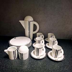 Op de afbeelding: Een wit porseleinen theeservies met zwarte en gouden accenten. De set bevat een theepot, borden, kopjes, schotels, een melkkan en een suikerpot. Het ontwerp heeft verticale zwarte strepen met gouden randen.