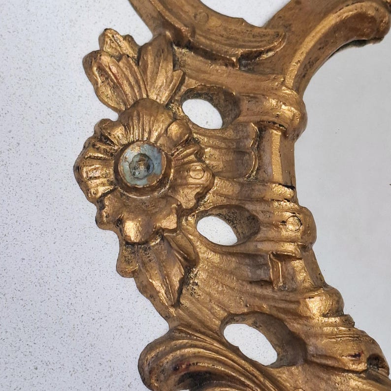 May include: Close-up of a gold-colored, ornate mirror frame. The frame features floral and scrollwork designs with a distressed finish. The mirror's edge is visible, reflecting a light-colored surface. The frame's details are highlighted by the lighting.