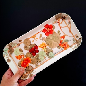 Peut inclure: Plateau rectangulaire avec un motif floral. Il présente des fleurs rouges, oranges et jaunes, des feuilles vertes et marron, et une bordure marron. Le texte "Tropaeolum Majus" est visible. Le fond est blanc.