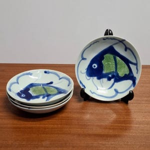 Op de afbeelding: Een set van vier kleine, witte keramische borden met een geschulpte rand. Elk bord heeft een blauw visontwerp met groene details. Eén bord wordt op een zwarte standaard getoond, de andere zijn opgestapeld. De borden staan op een houten oppervlak.