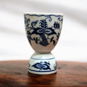 Könnte beinhalten: Ein weißer Eierbecher aus Keramik mit blauem Blumenmuster. Der Eierbecher hat eine abgerundete Oberseite und einen Sockel. Das blaue Design zeigt stilisierte Blumen und Blätter. Der Becher ist etwa 10 cm hoch.
