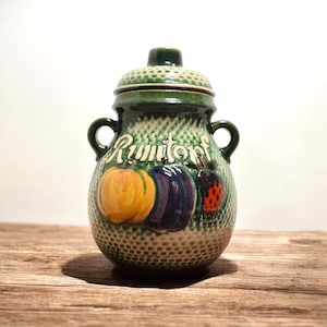 Op de afbeelding: Groene keramische Rumtopf-pot met een deksel, twee handvatten en een textuuroppervlak. De pot heeft geschilderde illustraties van een gele pompoen, een paarse pruim en een rode lieveheersbeestje. Het woord "Rumtopf" is in witte letters geschreven.