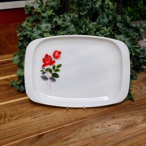 Op de afbeelding: Een witte rechthoekige serveerschaal met afgeronde hoeken. De schaal heeft een bloemmotief van twee rode rozen met groene bladeren en grijze stelen. De schaal wordt gepresenteerd op een houten oppervlak.