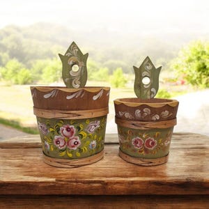 Op de afbeelding: Twee decoratieve houten emmers met bloemmotieven. De emmers zijn groen geschilderd met roze en witte bloemen. Elke emmer heeft een houten tulpvormige handgreep. De emmers staan op een houten oppervlak.