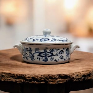 Puede incluir: Un cuenco de cerámica blanco con tapa y estampado floral azul. El cuenco tiene dos asas pequeñas y un pomo redondo en la tapa. El diseño presenta motivos florales y de hojas azules detallados.